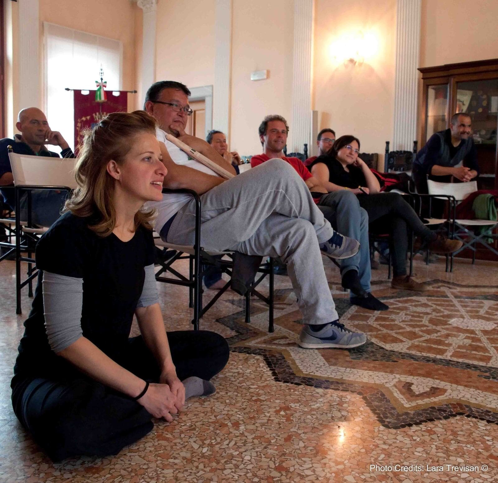 L'artista Caterina Comingio e un gruppo ristretto di persone, sono il pubblico di un laboratorio teatrale e guarda sorridendo davanti a sè.