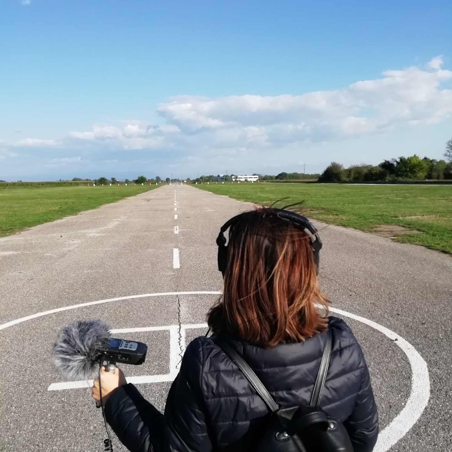 L'artista Carolina Valencia Caicedo in primo piano di spalle con cuffie alle orecchie e microfono in mano, è al centro di una pista di atterraggio di aerei.